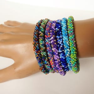 May include: A stack of colorful beaded bracelets in shades of green, blue, purple, and orange. The bracelets are made of small, round beads and are stacked on a person's wrist.