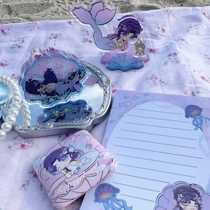 May include: A collection of mermaid-themed accessories and stationery. Includes a mirror, a small pink case, a notepad, and acrylic figurines. The items feature illustrations of mermaids, jellyfish, and fish in pastel colors, set on a floral patterned cloth.