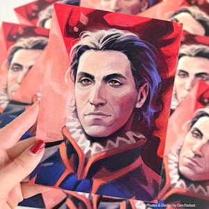 May include: A portrait print of a man with silver hair and a serious expression. The artwork uses a red and blue palette, with the man wearing a detailed collar and jacket. The print is held in a hand with red nail varnish.