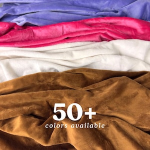 May include: Close-up of a stack of soft, plush fabric in various colors, including purple, pink, white, and brown. The text "50+ colors available" is printed on the brown fabric.