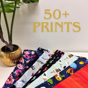 May include: A selection of fabric swatches with various patterns, including floral, superhero, and lightning bolt designs. The text "50+ PRINTS" is displayed in gold lettering. The fabrics are arranged in a fan shape.