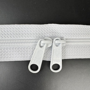May include: A close-up of a white zipper with two white plastic pull tabs. The pull tabs have the word "YKK" printed on them.