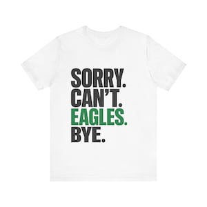 May include: A white short-sleeved t-shirt featuring the black text "SORRY. CAN'T." followed by the green text "EAGLES." and the black text "BYE." printed in a bold, sans-serif font centred on the front of the garment.