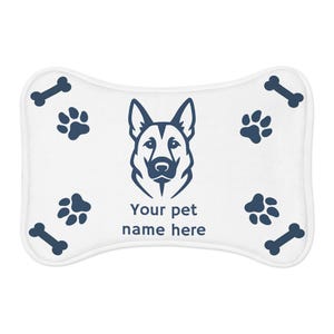 Puede incluir: Cama para mascotas en forma de hueso de perro blanco con un gráfico de pastor alemán azul marino. La cama presenta huellas de patas, huesos de perro y el texto "Your pet name here".