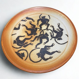 May include: A ceramic plate with a brown rim and a cream-colored center. The plate features a repeating pattern of black monkey figures within black circular designs. The monkeys are in various poses, creating a playful design.