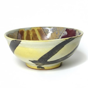 May include: A ceramic bowl with a cream-coloured exterior, accented by a bold black diagonal stripe and yellow and brown interior. The bowl has a rounded shape and a slightly flared rim, showcasing a unique artistic design.