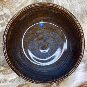 May include: A brown ceramic bowl with a blue glaze. The bowl has a swirl pattern and is glazed on the inside and outside.