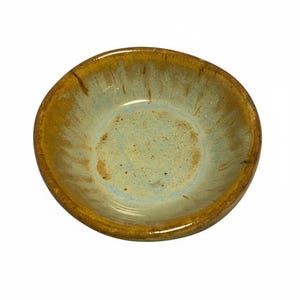 May include: A small, handmade ceramic bowl with a unique glaze. The bowl features a light blue and tan interior with a darker brown rim. The glaze has a speckled appearance, adding to its rustic charm. The bowl is perfect for serving small portions.