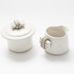 Peut inclure: Un sucrier en céramique blanche avec couvercle et une petite crémière. Les deux pièces ont une finition mouchetée et sont ornées d'une poignée unique. Les poignées et le bouton du couvercle présentent un faisceau de tiges blanches enveloppées d'une bande beige.