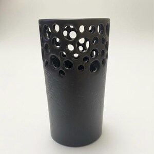 May include: A dark brown ceramic vase with a textured surface. The upper portion features a pattern of varying sized circular cutouts. The vase has a cylindrical shape and stands on a white background.