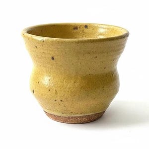 May include: A small, handmade ceramic cup with a yellow-ochre glaze. The cup has a rounded body and a slightly flared rim. The base is a darker, unglazed brown. The surface has a textured appearance with small, dark speckles.