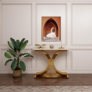 May include: A modern console table with a marble top and gold-colored curved legs. A framed print of an arched hallway hangs above the table. A potted plant with large green leaves and a woven basket sits to the left.