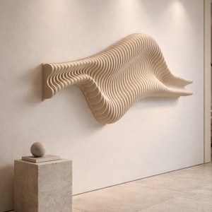 May include: A light-colored, wave-shaped wall sculpture made of layered wood. The artwork is mounted on a white wall. A small, square stone pedestal with a sphere on top is in the foreground.