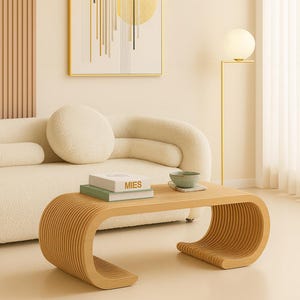 Può includere: Un tavolino da caffè in legno chiaro con un design curvo e a doghe. Il tavolo ha un piano rettangolare con bordi arrotondati e una base curva. Due libri e una piccola ciotola sono sul tavolo. La parola "MIES" è visibile sul libro.