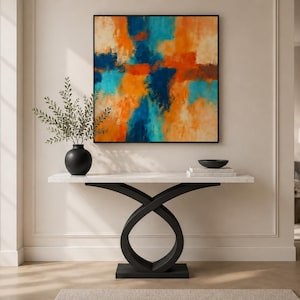 Könnte beinhalten: Ein abstraktes Gemälde in einem schwarzen Rahmen hängt über einem weißen Marmor-Konsolentisch mit schwarzem Sockel. Das Gemälde zeigt orange, blaue und cremefarbene Farben. Eine schwarze Vase mit Grün und eine schwarze Schale stehen auf dem Tisch.