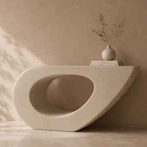 May include: A contemporary, cream-coloured console table with a distinctive, curved form. The table has a large, oval aperture and a layered, textured surface. A small, round vase with greenery and books are on top.