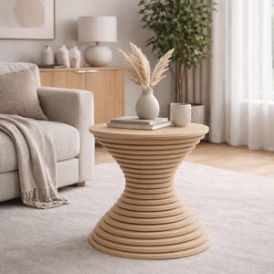 May include: A light brown, spiral-shaped side table with a vase of dried flowers, a stack of books, and a white mug. The table is in a living room setting with a beige sofa, a wooden cabinet, and a large potted plant. The table is approximately 24 inches tall.