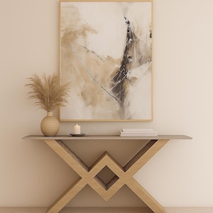 May include: A contemporary console table with a light grey surface and a geometric wooden base. The table holds a vase with dried plants, a lit candle, and books. An abstract painting in a gold frame hangs on the wall.