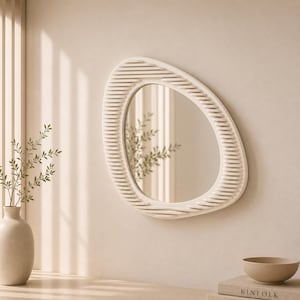 Op de afbeelding: Een off-white, ovaalvormige wandspiegel met een getextureerde, geribbelde lijst. De spiegel is gemonteerd op een neutraal gekleurde muur en weerspiegelt een glimp van groen. Het ontwerp is modern en minimalistisch, geschikt voor woondecoratie.