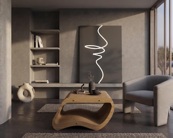 May include: A living room interior featuring a light wood coffee table with a curved, layered base, a gray upholstered armchair, and a large rectangular wall art piece displaying a single continuous white glowing line on a dark gray background.