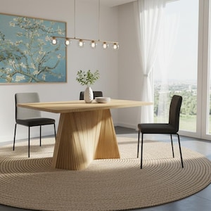 May include: A modern dining room featuring a light wood table with a unique geometric base, surrounded by black chairs. A large, round woven rug sits beneath the table. A framed floral painting hangs on the wall, and a window with white curtains is visible.