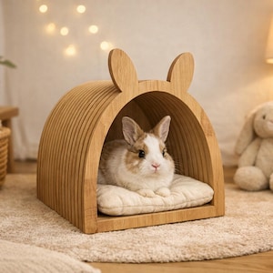 May include: A wooden rabbit house with a rounded top and bunny ear accents. A small, brown and white rabbit rests on a cream-colored cushion inside the house. The house is on a beige rug, with a stuffed bunny and a lamp in the background.