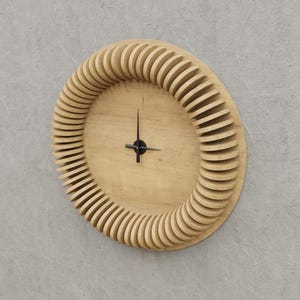 Op de afbeelding: Een houten wandklok met een uniek ontwerp met een ronde lijst gemaakt van gelaagde, overlappende houten stukken. De wijzerplaat van de klok is eenvoudig en heeft zwarte wijzers.