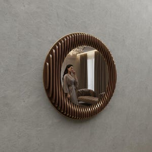 May include: A round decorative mirror with a brown wooden frame and a gold-colored trim. The frame has a unique, layered design. The mirror reflects a room with a person in a robe.