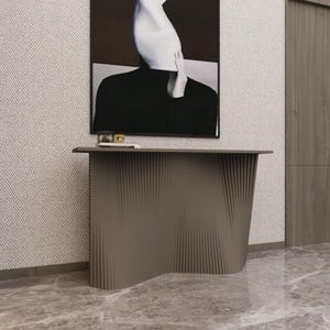 May include: A contemporary, taupe console table with a distinctive, undulating design. The table is placed in front of a wall featuring a black and white artwork and a wooden door. The floor is polished marble.