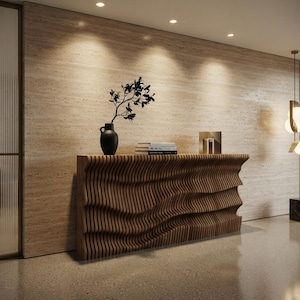 May include: A long console table constructed from vertical wooden slats arranged in a flowing, wave-like pattern. A matte black vase containing a leafy branch and a small stack of books are placed on the flat top surface of the wooden furniture piece.