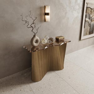 May include: A console table with a wavy, transparent glass top and a fluted, light brown wooden base. On the table sits a beige, donut-shaped vase with twisted branches, three clear glass spheres, and a small rectangular wooden box with a gold latch.