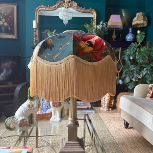 May include: A vintage table lamp with a floral patterned lampshade in teal and red, trimmed with gold and long, tan fringe. The lamp has a brass base and a square foot. The lamp is on a glass table.