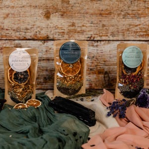 May include: Three clear pouches filled with dried herbs, spices, and citrus slices. Each pouch has a circular label with text. The pouches are arranged on a white and green fabric with dried lavender and a black stone.