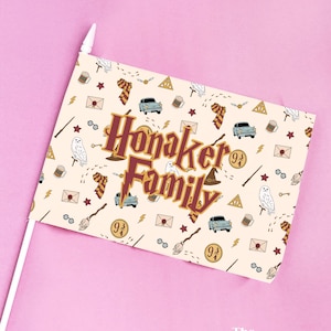 May include: A rectangular flag with a white pole, featuring a cream-colored background with repeating Harry Potter-themed illustrations. The flag displays the text "Honaker Family" in a burgundy and gold font, with a pink background.