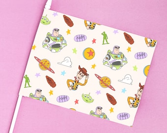 Puede incluir: Una bandera con un patrón repetido de personajes de Toy Story, incluyendo a Woody, Buzz Lightyear y los pequeños hombres verdes, sobre un fondo crema. La bandera está montada en un palo blanco sobre un fondo rosa.