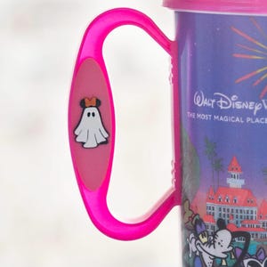 Può includere: Una tazza da viaggio viola con coperchio e manico rosa acceso. Il manico presenta una piccola icona di un fantasma con orecchie arancioni. La tazza mostra una grafica di un edificio e personaggi Disney con il testo Walt Disney World The Most Magical Place.