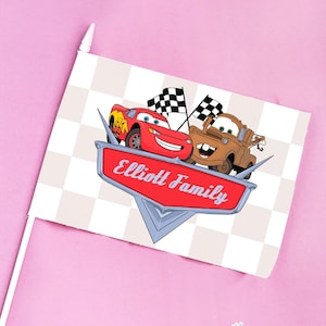 May include: A small flag featuring Lightning McQueen and Mater from the Cars film, with the text "Elliott Family" on a red emblem. The background is a checkered pattern, and the flag is on a white stick.