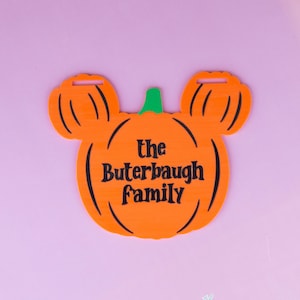 Puede incluir: Un letrero naranja con forma de calabaza, un tallo verde y dos orejas más pequeñas con forma de calabaza en la parte superior. El texto "the Buterbaugh family" está impreso en negro en el centro. Tiene dos ranuras rectangulares arriba.
