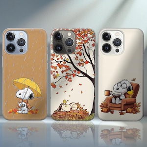 May include: Three phone cases with Snoopy designs in autumnal settings. One depicts Snoopy with an umbrella in the rain, another with Snoopy asleep in leaves, and the last with Snoopy relaxing in a chair with a coffee cup.