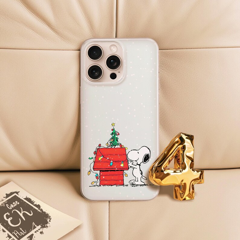 May include: A clear phone case featuring a Snoopy Christmas design with a red doghouse, a decorated Christmas tree, and Snoopy. A gold number 4 and a business card are also in the image.