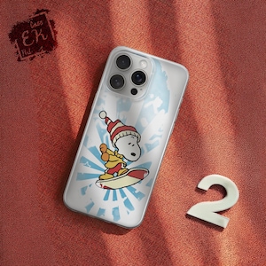 May include: Clear phone case featuring a cartoon Snoopy character wearing a Santa hat and surfing on a red and white surfboard. The design includes a blue and white sunburst background. The case is on a red-orange textured surface.
