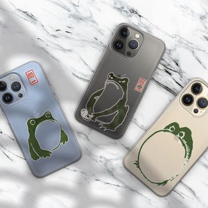 Puede incluir: Tres fundas para teléfono con diseños de ranas. Una es azul claro, otra gris y otra beige. Cada funda tiene una ilustración de rana verde y un cuadrado rojo con texto japonés. Las fundas están sobre una superficie de mármol.