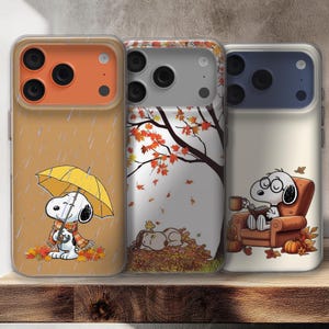Puede incluir: Tres fundas para teléfono con diseños de Snoopy en escenas otoñales. Una funda muestra a Snoopy con un paraguas, otra a Snoopy durmiendo entre hojas y la tercera a Snoopy leyendo. Las fundas tienen carcasas de cámara naranja, plateada y azul.