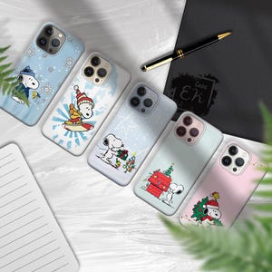 May include: A collection of phone cases featuring Snoopy in various winter and holiday scenes. The cases showcase Snoopy with snowflakes, surfing, and decorating a Christmas tree. The cases are in shades of blue, white, and pink.