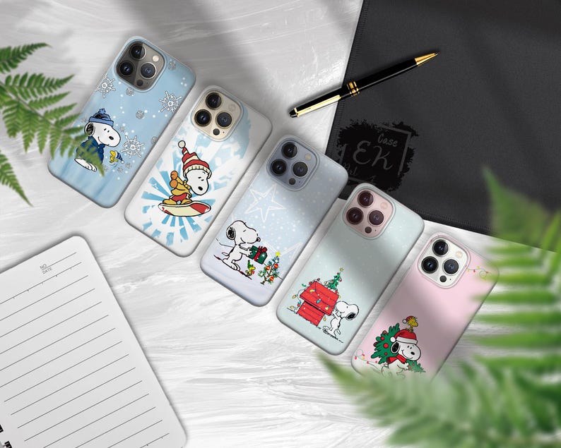 May include: A collection of phone cases featuring Snoopy in various winter and holiday scenes. The cases showcase Snoopy with snowflakes, surfing, and decorating a Christmas tree. The cases are in shades of blue, white, and pink.