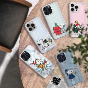 May include: A collection of phone cases featuring Snoopy in various winter and Christmas scenes. The cases are white, light blue, and pink, with Snoopy wearing a Santa hat, holding gifts, and snowboarding. The cases have a glossy finish.