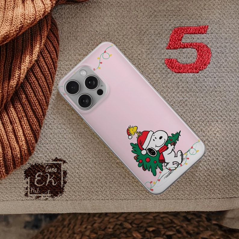 May include: A pink phone case featuring a Christmas-themed Snoopy and Woodstock design. Snoopy is wearing a Santa hat and holding a Christmas tree. The case has a clear border with decorative lights.