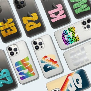 May include: A selection of clear phone cases featuring diverse, colourful graphic designs. Designs include glitter letters, rainbow patterns, and retro-style graphics. The cases are arranged on a light blue surface.