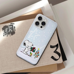 May include: A light blue iPhone case featuring a Snoopy graphic holding a gift box near a Christmas tree. The case has a star and snow design. The case is on top of a stack of books.