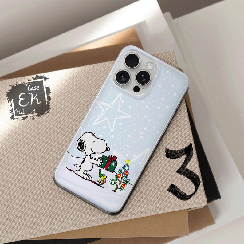 May include: A light blue iPhone case featuring a Snoopy graphic holding a gift box near a Christmas tree. The case has a star and snow design. The case is on top of a stack of books.